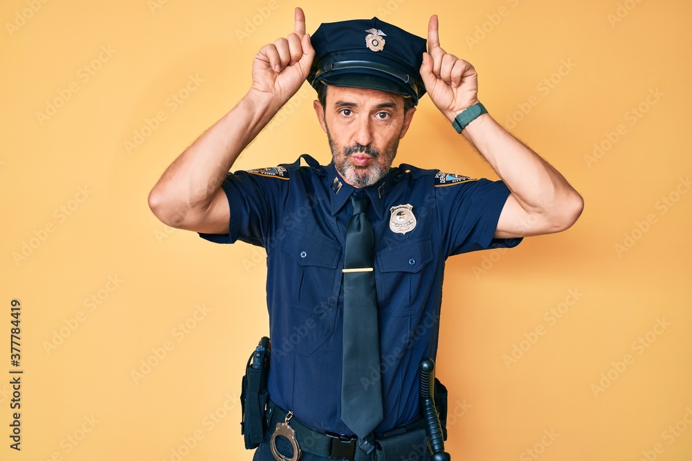 Middle age hispanic man wearing police uniform doing funny gesture with finger over head as bull horns Middle age hispanic man wearing police uniform doing funny gesture with finger over head as bull horns