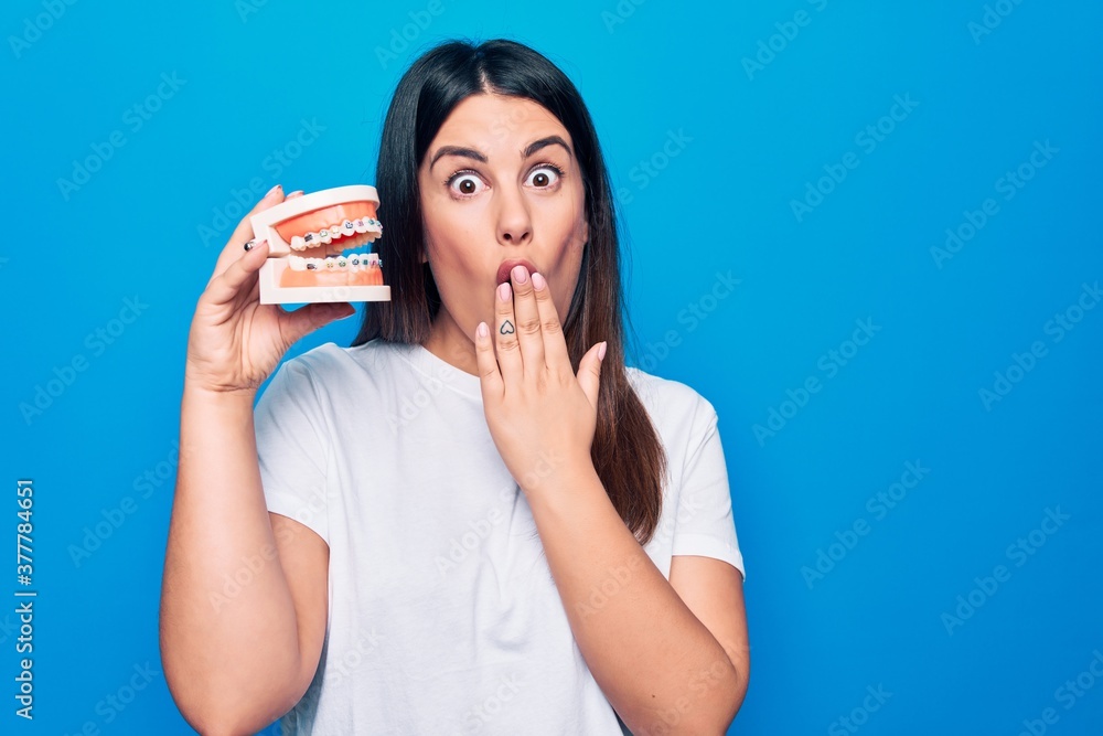 Young beautiful brunette woman asking for dental care holding denture with backets covering mouth with hand, shocked and afraid for mistake. Surprised expression Young beautiful brunette woman asking for dental care holding denture with backets covering mouth with hand, shocked and afraid for mistake. Surprised expression