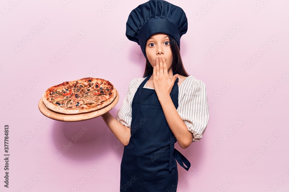 Beautiful child girl wearing cooker uniform holding italian pizza covering mouth with hand, shocked and afraid for mistake. surprised expression Beautiful child girl wearing cooker uniform holding italian pizza covering mouth with hand, shocked and afraid for mistake. surprised expression