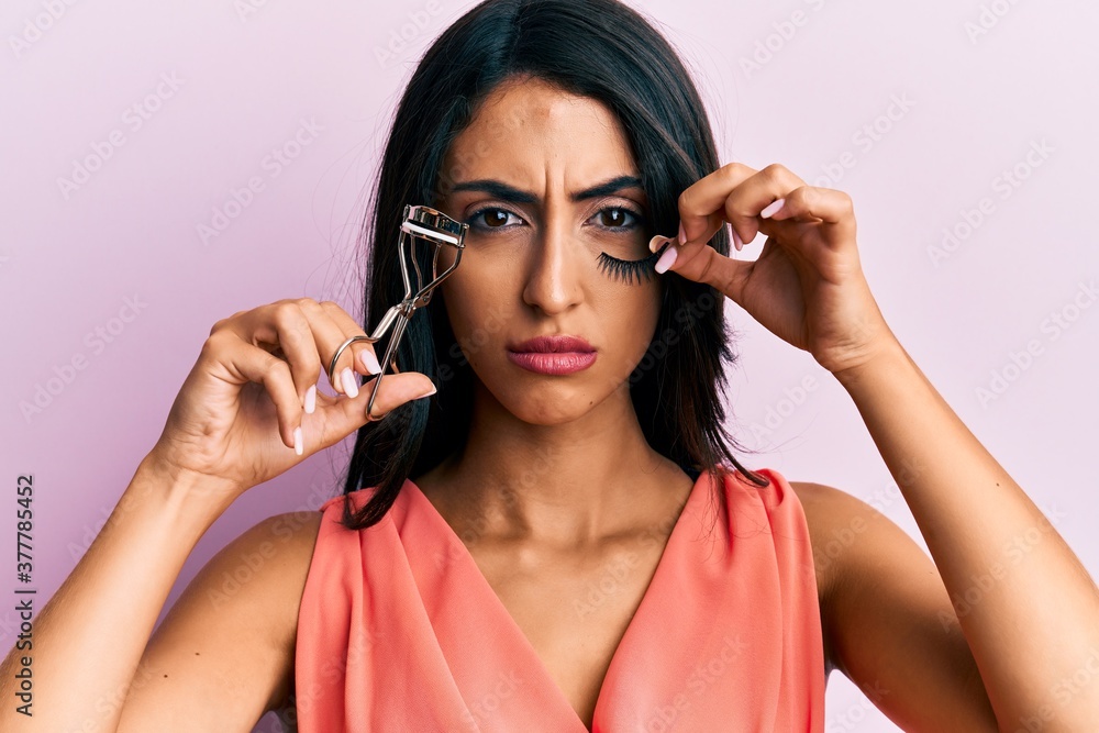 Beautiful hispanic woman holding eyelash curler and fake lashes skeptic and nervous, frowning upset because of problem. negative person. Beautiful hispanic woman holding eyelash curler and fake lashes skeptic and nervous, frowning upset because of problem. negative person.