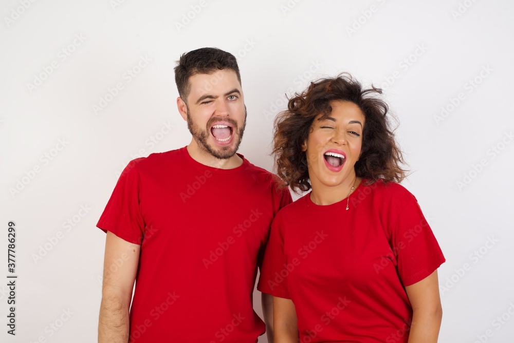 Young beautiful couple wearing red t-shirt on white background winking looking at the camera with sexy expression, cheerful and happy face. Young beautiful couple wearing red t-shirt on white background winking looking at the camera with sexy expression, cheerful and happy face.
