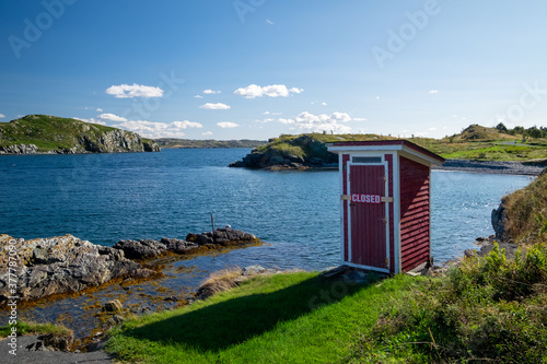 Foto A small red building, outhouse, on the edge of a rocky cliff with blue ocean and islands in the background