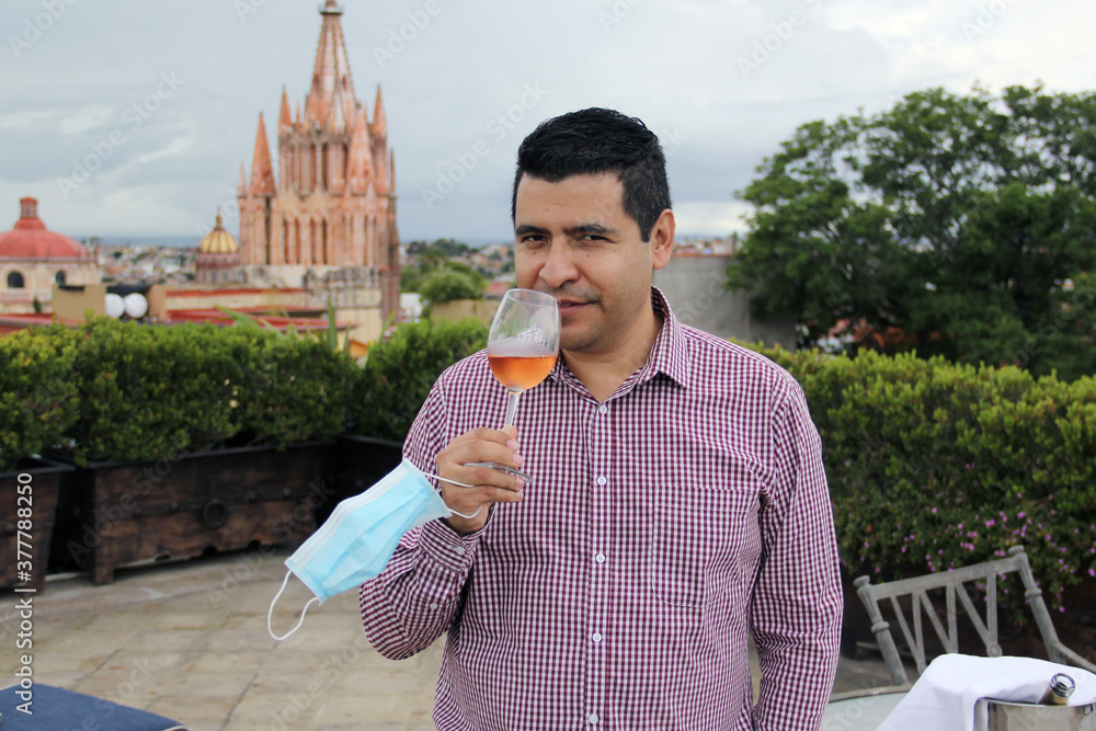 Fototapeta premium Casual latin man with protection mask, tasting rosé sparkling wine in crystal goblet in outdoor terrace with view of the cathedral in San Miguel de Allende Guanajuato Mexico, new normal