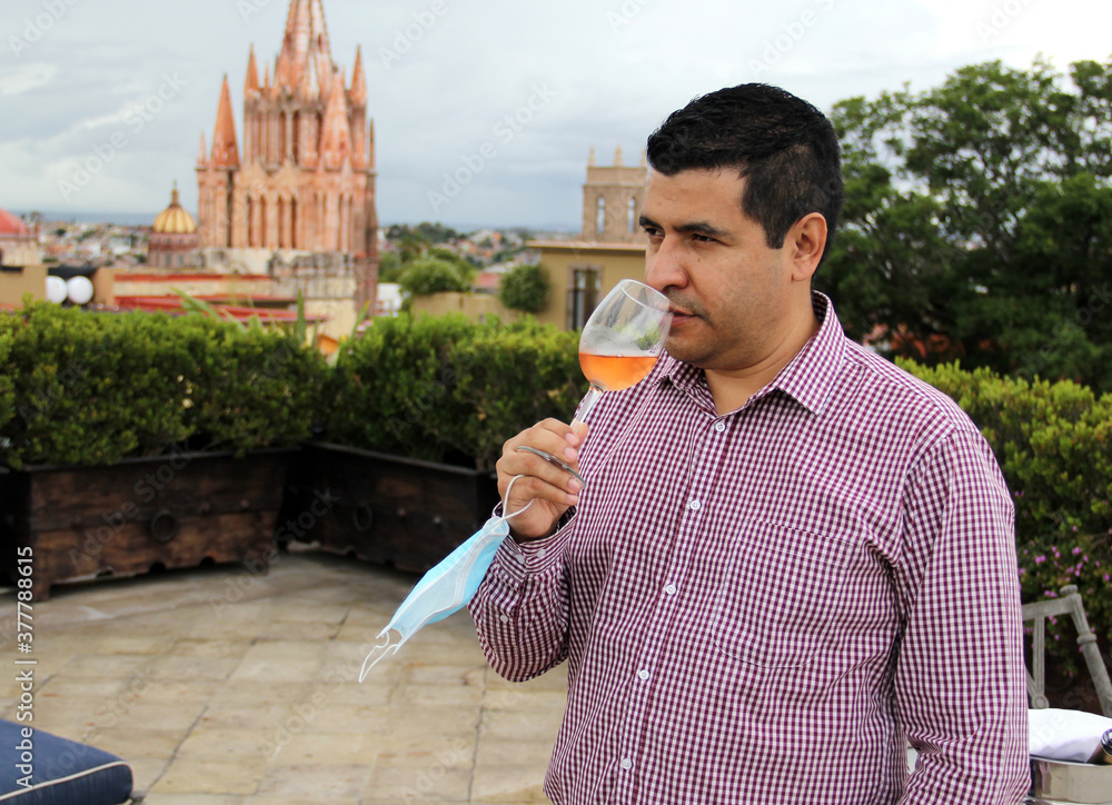 Fototapeta premium Casual latin man with protection mask, tasting rosé sparkling wine in crystal goblet in outdoor terrace with view of the cathedral in San Miguel de Allende Guanajuato Mexico, new normal