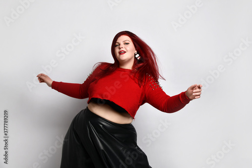 Obese ginger lady in red spiked top, black bra and leather skirt. She is dancing, posing isolated on white photo background