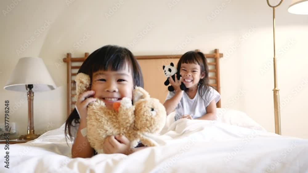Two little sibling girls in white pajamas are hugging a doll lying on bed together in the bedroom. Concept of Childhood happiness. Two little sibling girls in white pajamas are hugging a doll lying on bed together in the bedroom. Concept of Childhood happiness.
