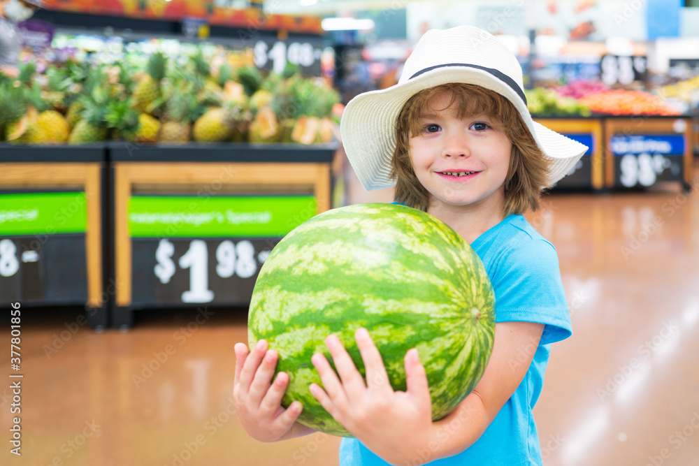 Fototapeta premium Kid holding watermelon in supermarket against food stall. Smiling positive boy grocery shopping at the supermarket.