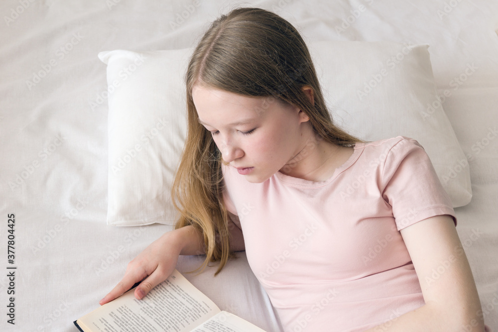 A young girl with long blonde hair is reading a book while lying on the bed A young girl with long blonde hair is reading a book while lying on the bed