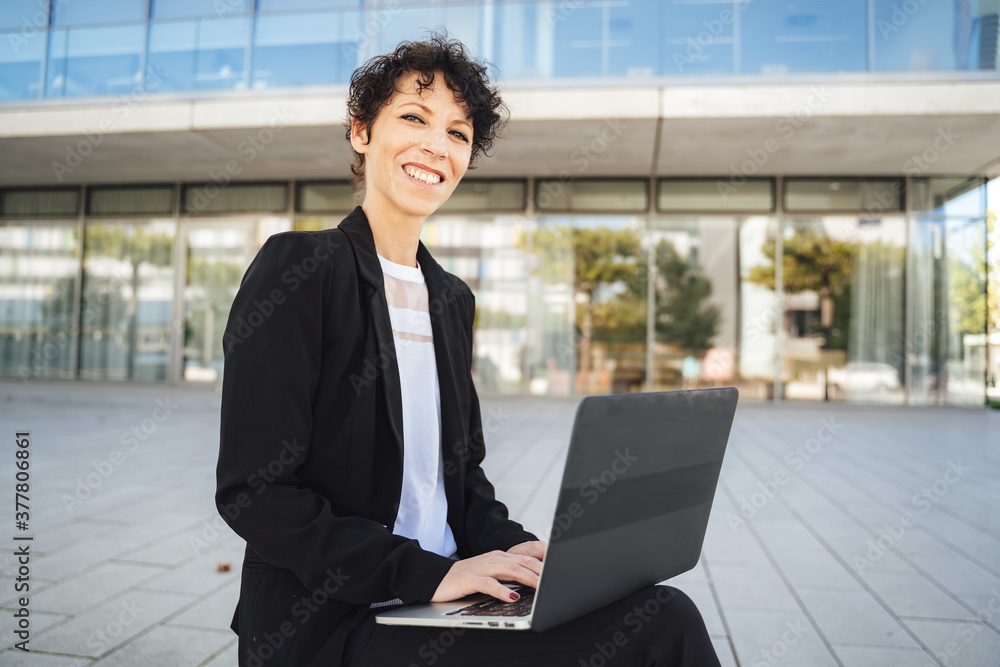 Portrait of smiling mature businesswoman in suit using laptop while sitting outside office Portrait of smiling mature businesswoman in suit using laptop while sitting outside office