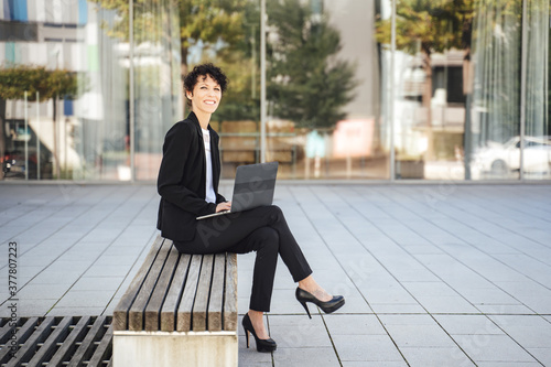 Full length portrait of smiling mature businesswoman with laptop sitting on bench outside office in city