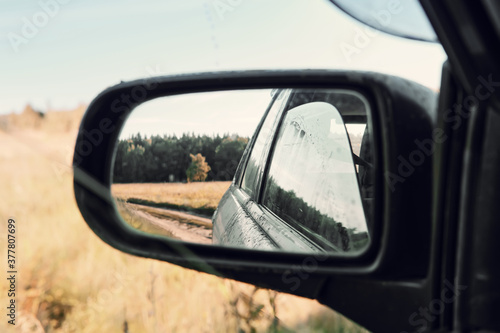 Reflection of dirty car and landscape in left rearview mirror on sunny summer day