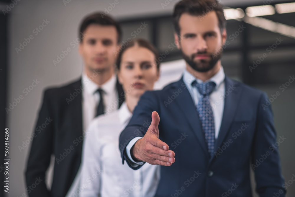 Bearded male showing outstretched hand, his female and male collegues standing behind