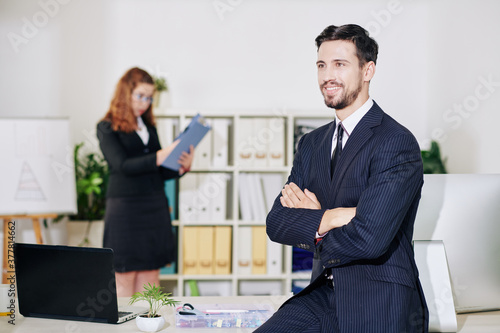 Smiling young businessman sitting on table with arms folded and looking away