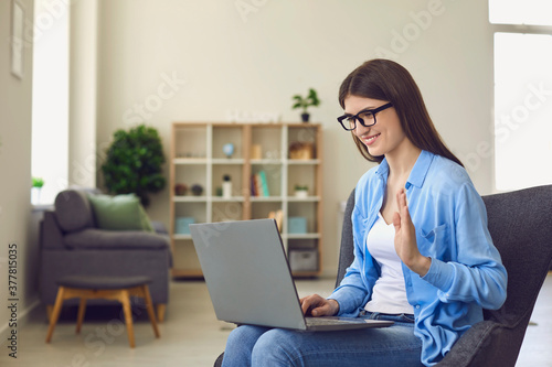 Young woman in glasses making video call on laptop and waving hand to greet relatives or colleagues