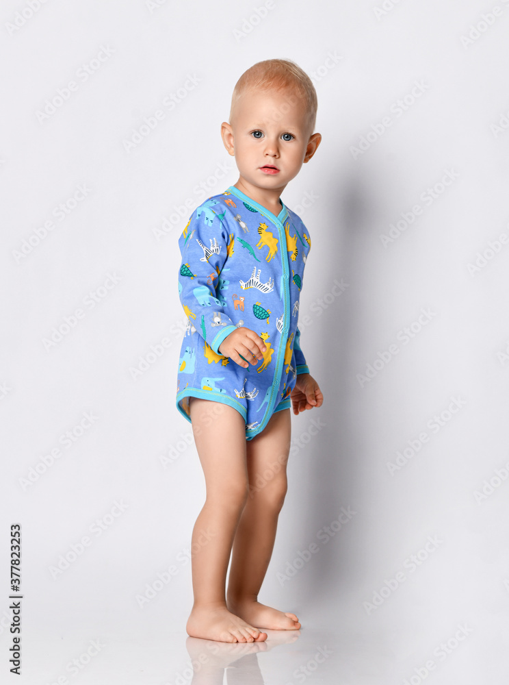 Baby boy in blue one-piece bodysuit romper with long sleeves stands side looking at camera Baby boy in blue one-piece bodysuit romper with long sleeves stands side looking at camera