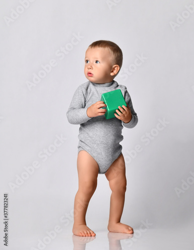 Toddler one-year-old baby boy in diaper and grey one-piece bodysuit with long sleeves stands holding green toy block