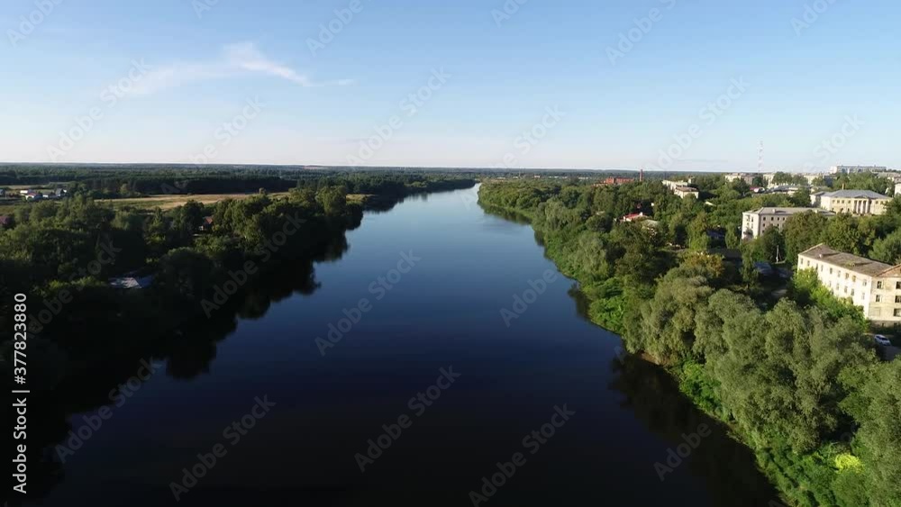 Top view of a wide river with blue water. There are dense green trees along the shore. On the side is the outskirts of the city. Clear sky over the reservoir. Smooth video motion. Summer sunny day. Top view of a wide river with blue water. There are dense green trees along the shore. On the side is the outskirts of the city. Clear sky over the reservoir. Smooth video motion. Summer sunny day.