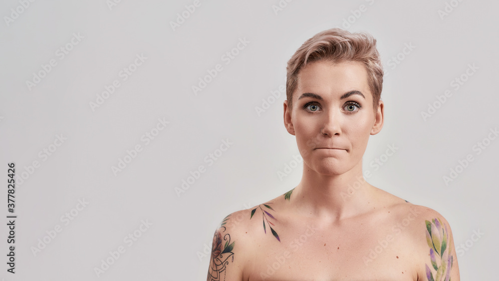 Obraz premium Portrait of a young attractive half naked tattooed woman bites and presses lips in bewilderment, reacts on something, looking at camera isolated over light background