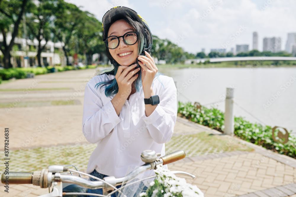 Happy beautiful young Chinese woman with blue hair putting on helmet before riding bicycle around city pond