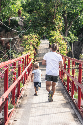 Back view Father son in t-shirt running across red iron suspension bridge way to forest park. Friendly family vacation outdoor active enjoying leisure time nature concept. Run away from the camera