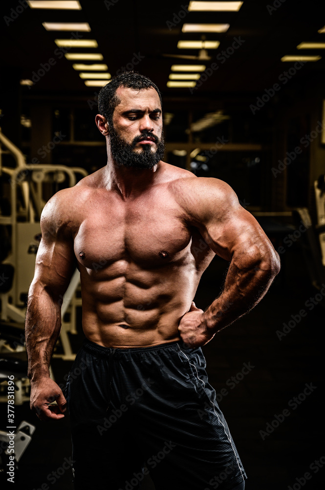handsome bearded young athlete man with strong muscular physique body in dark sport gym