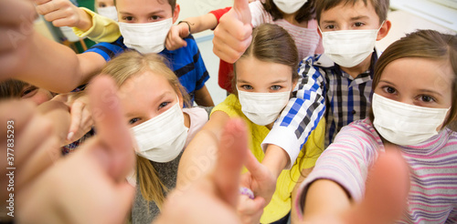 education, people and pandemic concept - group of children wearing face protective medical mask for protection from virus disease showing thumbs up at school