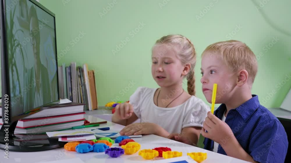 boy with Down syndrome at home education with sister listen to the female teacher from the computer screen sitting at desk with cards in room boy with Down syndrome at home education with sister listen to the female teacher from the computer screen sitting at desk with cards in room