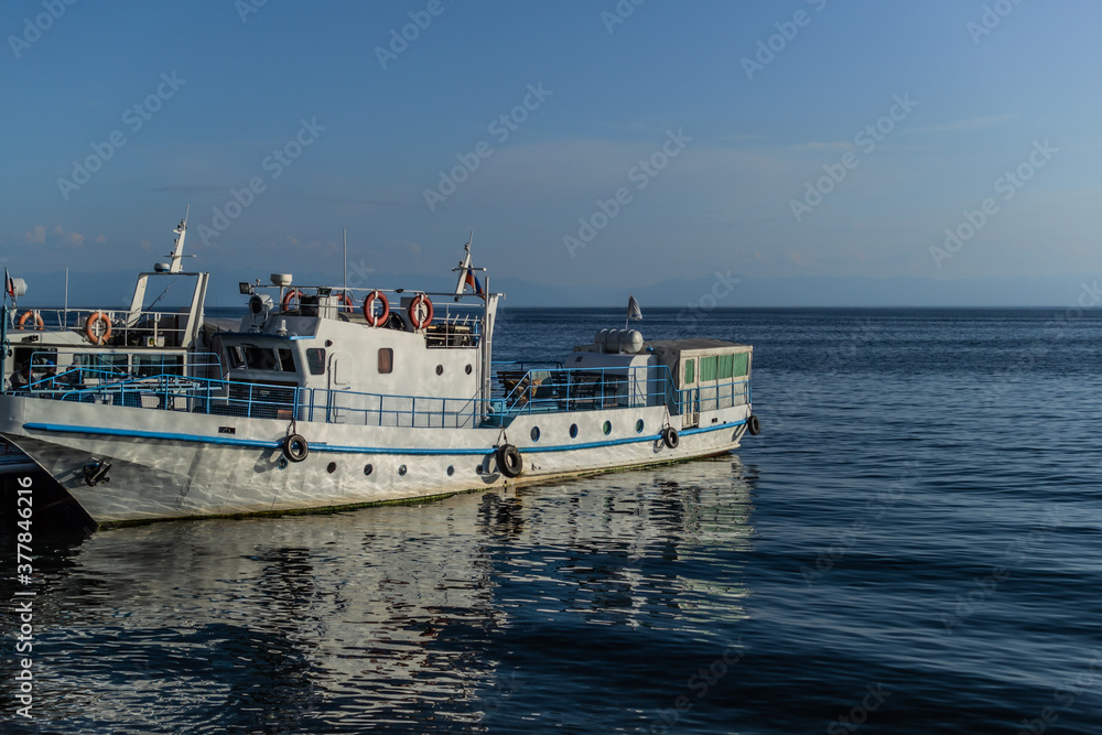 white vessel ship boat with blue stripes on pier in bay of lake baikal in the light of the sun with reflections in water, horizon