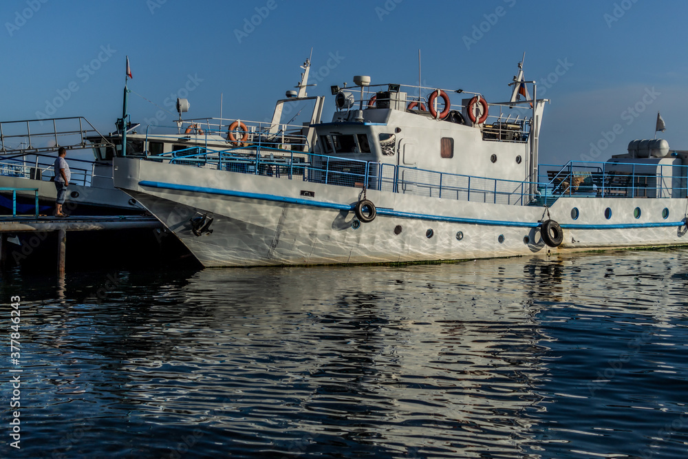white ship boat with blue stripes on the pier in bay of lake Baikal in the light of sun with reflections