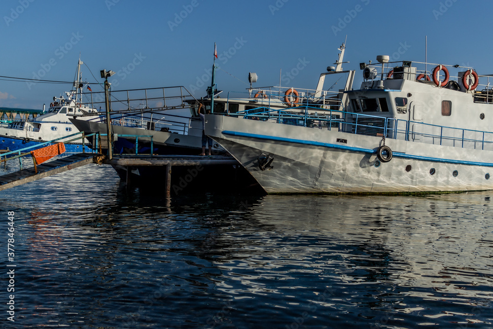 Naklejka premium white vessel ship boat with blue stripes on pier in bay of lake baikal in light of sun with reflections in water