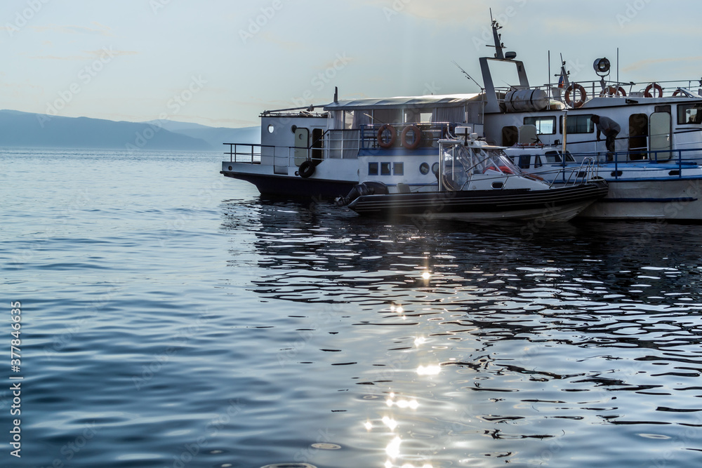 Naklejka premium white ships boats on pier in bay of blue lake baikal with glowing and reflections in water in light of sunset