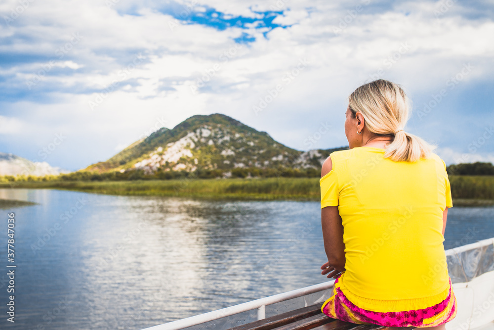 Fototapeta premium A blonde girl on the river bank looks into the distance at a mountain landscape - a view from the back in a yellow T-shirt