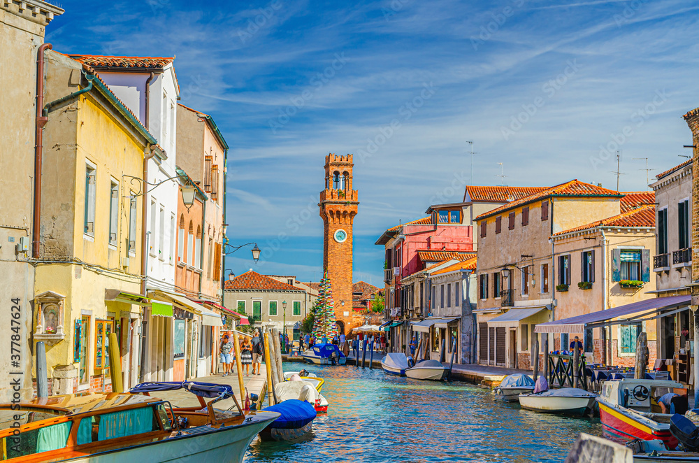 Murano islands with clock tower Torre dell'Orologio, boats and motor boats in water canal, colorful traditional buildings, Venetian Lagoon, Veneto Region, Northern Italy. Murano postcard cityscape.