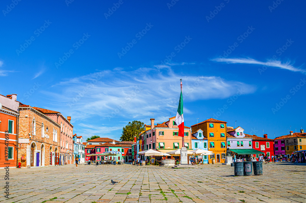 Burano island central town square with old colorful multicolored buildings and italian flag, blue sky in sunny summer day background, Venice Province, Veneto Region, Northern Italy. Burano postcard