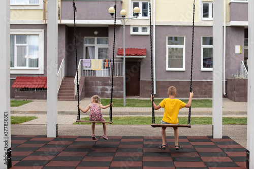 A 7-year-old blond boy in a bright yellow t-shirt and a 4-year-old blonde girl ride a swing on a Playground in the courtyard of a multi-storey building