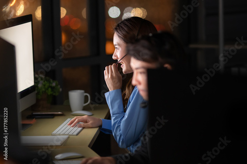 Young and beautiful call center officers team work overtime with happy at 24/7 hour at night with bokeh of light in background. Concept for customer service late working for support as back office