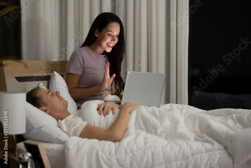 Woman housewife sitting on bed in the night bedtime and using laptop notebook computer connect to internet while husband sleeping beside her. She greeting and talk to someone in social media.