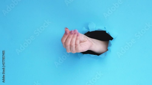 Pay back time. Debt loan. Female hand asking for money with gesture inside breakthrough hole isolated on blue ripped paper wall background with copy space. Commercial banner.