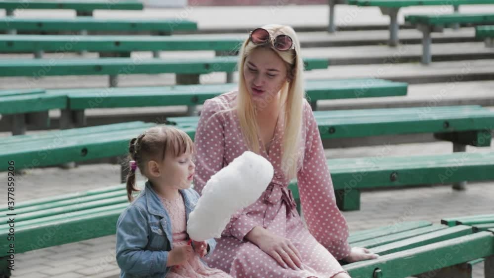 Beautiful and happy little girl eating cotton candy on the bench. Young happy mother with little girl. Beautiful and happy little girl eating cotton candy on the bench. Young happy mother with little girl.