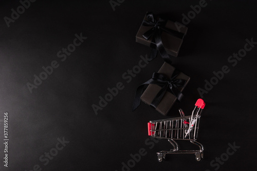 Black Friday sale shopping concept, Top view of gift box wrapped black paper and black bow ribbon present dropping to the shopping cart, studio shot on black background