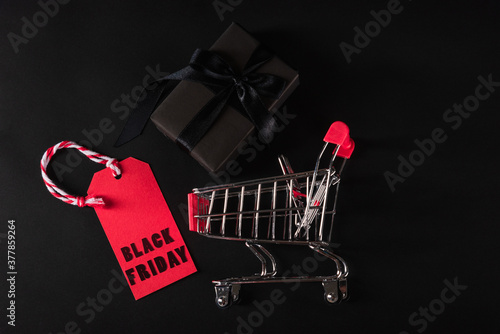 Black Friday sale shopping concept, Top view of gift box wrapped black paper and black bow ribbon present in the shopping cart and red tag label, studio shot on black background