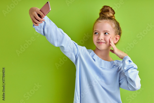 sweet positive child girl take selfie on mobile phone, take photo of herself, looking at screen of smartphone, isolated over green background