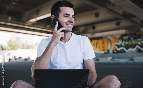 Pleased man speaking on smartphone while sitting on street with laptop