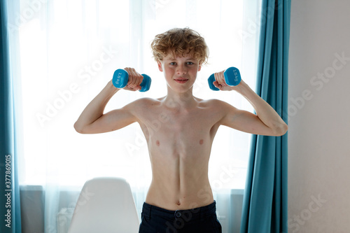 shirtless young caucasian strong teenager boy exercising at home, hold dumbbells in hands, pumping arm muscles. sport concept photo