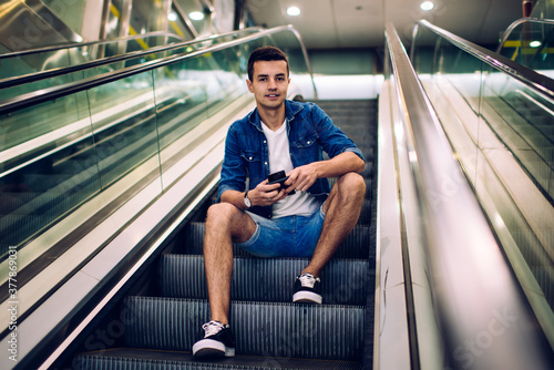 Young man sitting on steps of escalator with mobile phone