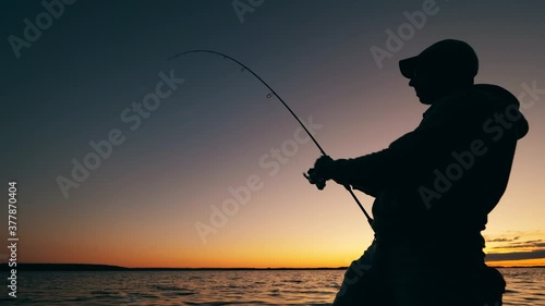 A man is reeling up a fishing pole in a sunset lake