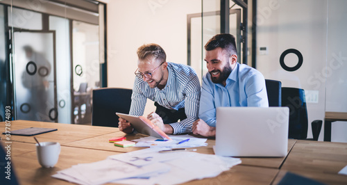 Successful young male colleagues having friendly conversation while making research for cooperation project, smiling happy businessmen laughing and having fun during working process in office