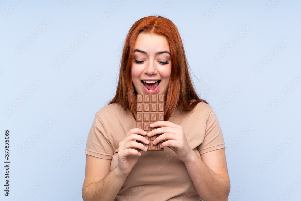 Redhead teenager girl over isolated blue background eating a chocolate tablet Redhead teenager girl over isolated blue background eating a chocolate tablet