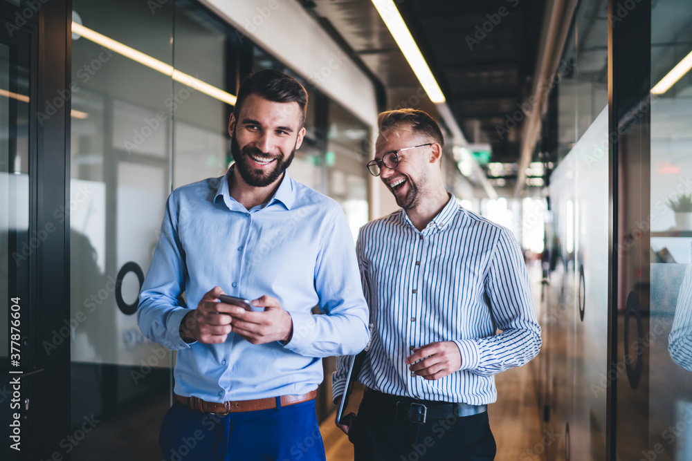 Cheerful male colleagues enjoying partnership in friendly working environment walking in office hallway and laughing while discussing web publication from smartphone, happy employees satisfied Cheerful male colleagues enjoying partnership in friendly working environment walking in office hallway and laughing while discussing web publication from smartphone, happy employees satisfied