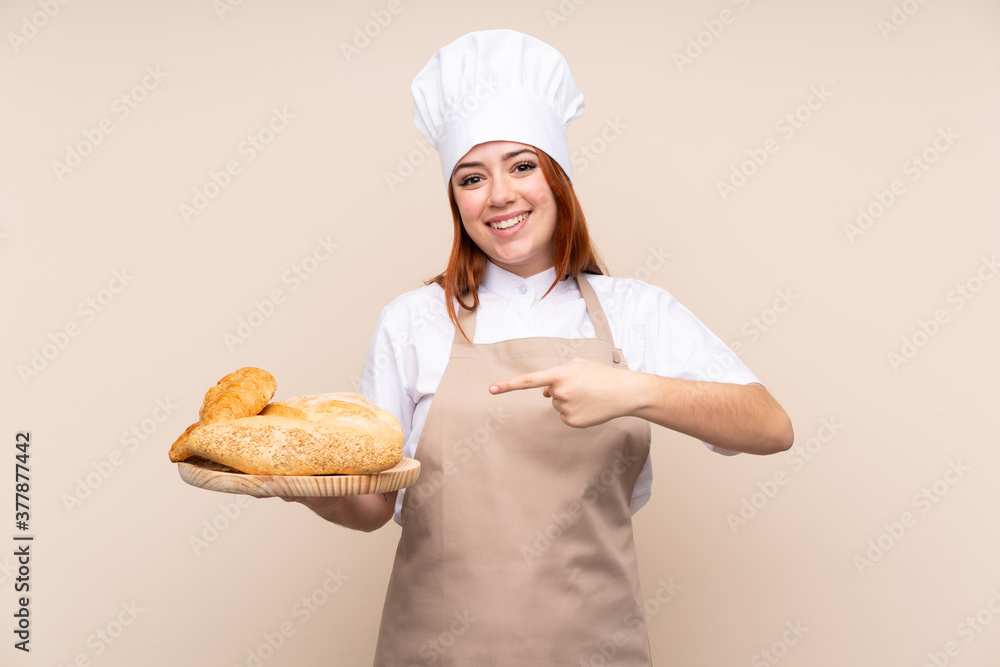 Redhead teenager girl in chef uniform. Female baker holding a table with several breads and pointing it Redhead teenager girl in chef uniform. Female baker holding a table with several breads and pointing it